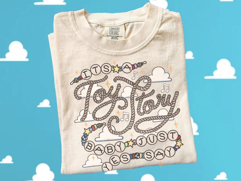 May include: A cream-colored short-sleeve t-shirt featuring a graphic design with the text 'ITS A Toy Story BABY JUST YES SAY'. The lettering is styled like rope, surrounded by colorful beads, stars, musical notes, and cloud illustrations.