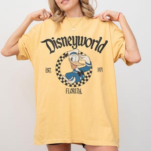 May include: Yellow t-shirt with a Disneyworld graphic featuring Donald Duck in a sailor outfit. The design includes the text "Disneyworld", "EST. 1971", and "FLORIDA" in black lettering. The shirt has a relaxed fit.