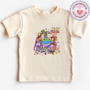 May include: A cream-colored short-sleeve t-shirt featuring a graphic print of Toy Story 5 characters including Woody, Buzz Lightyear, and others emerging from a purple backpack, with the text Toy Story 5 printed in blue and yellow above the characters.