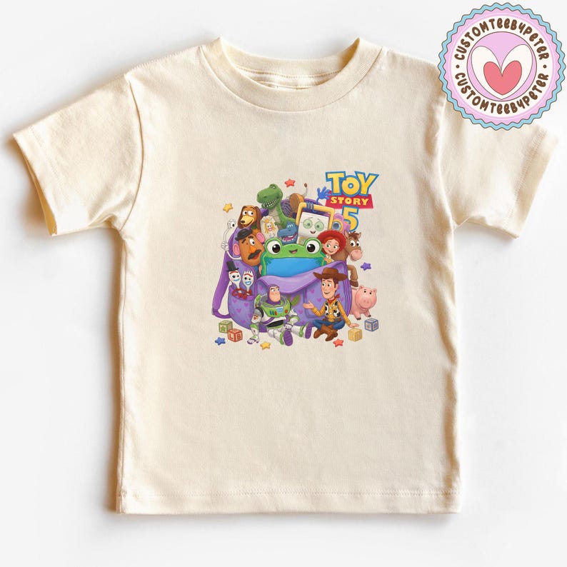 May include: A cream-colored short-sleeve t-shirt featuring a graphic print of Toy Story 5 characters including Woody, Buzz Lightyear, and others emerging from a purple backpack, with the text Toy Story 5 printed in blue and yellow above the characters.