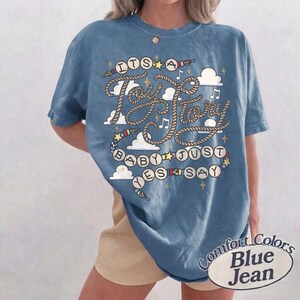 May include: A blue short-sleeve t-shirt featuring a graphic print with the text "ITS A Toy Story BABY JUST YES SAY". The design includes rope-style lettering, white clouds, small stars, musical notes, and colorful beads on a string.