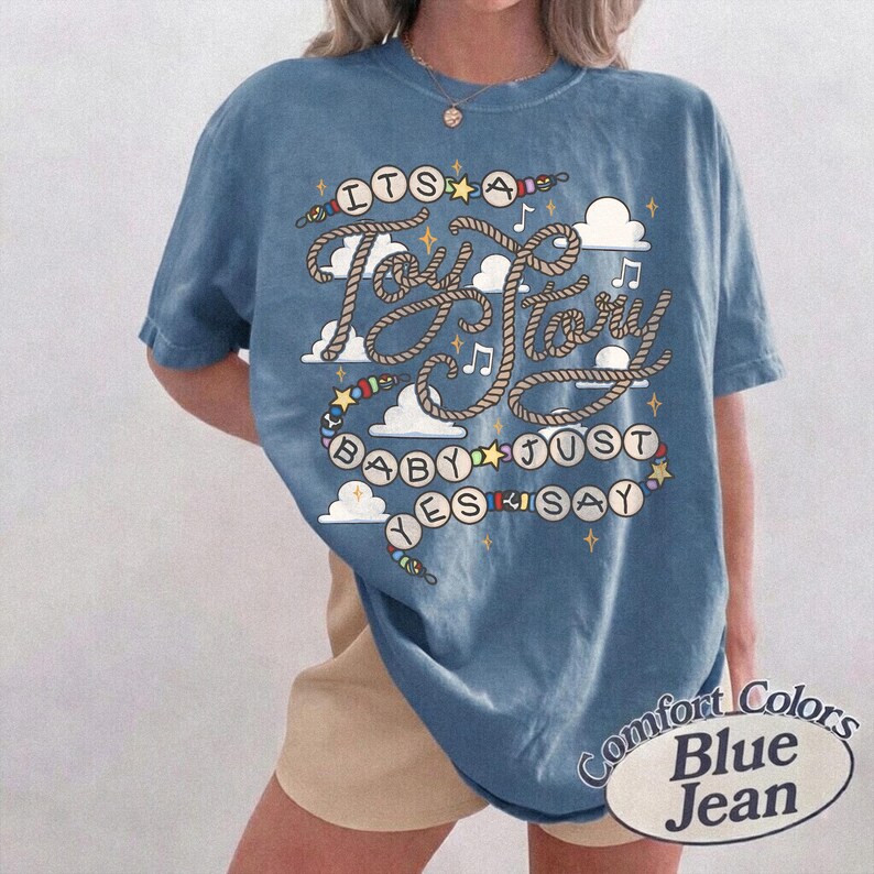 May include: A blue short-sleeve t-shirt featuring a graphic print with the text "ITS A Toy Story BABY JUST YES SAY". The design includes rope-style lettering, white clouds, small stars, musical notes, and colorful beads on a string.