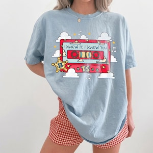 May include: A light blue t-shirt featuring a graphic of a red cassette tape surrounded by white clouds, yellow stars, and musical notes. The cassette tape displays the text "I KNEW IT, I KNEW YOU" and "TS" with a yellow star icon labeled "13".