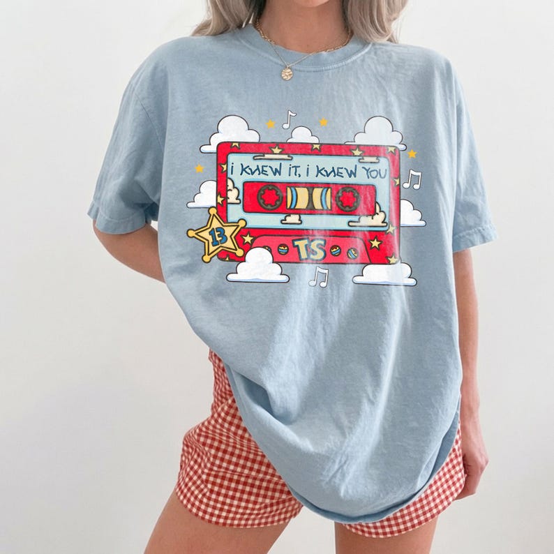 May include: A light blue t-shirt featuring a graphic of a red cassette tape surrounded by white clouds, yellow stars, and musical notes. The cassette tape displays the text "I KNEW IT, I KNEW YOU" and "TS" with a yellow star icon labeled "13".