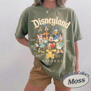 K&ouml;nnte beinhalten: Ein olivgr&uuml;nes Kurzarm-T-Shirt mit einer Grafik von Mickey Maus, Minnie Maus, Donald Duck, Daisy Duck, Goofy und Pluto. Der Text lautet Disneyland, Est. 1955, California, und enth&auml;lt die Namen der Figuren in einer Schreibschrift.