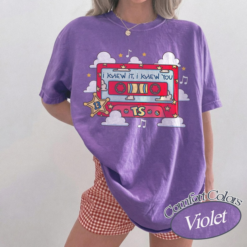 May include: A violet short-sleeved t-shirt featuring a graphic of a red cassette tape with the text "I KNEW IT, I KNEW YOU" and "TS". The design includes white clouds, yellow stars, and musical notes surrounding the cassette tape illustration.