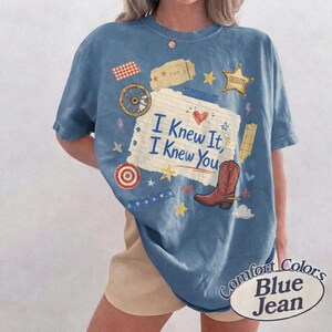 May include: A blue short-sleeve t-shirt featuring a graphic print of a lined notebook paper with the text "I Knew It, I Knew You" in blue script. The design includes a red cowboy boot, a sheriff star, a wagon wheel, and various small stars.