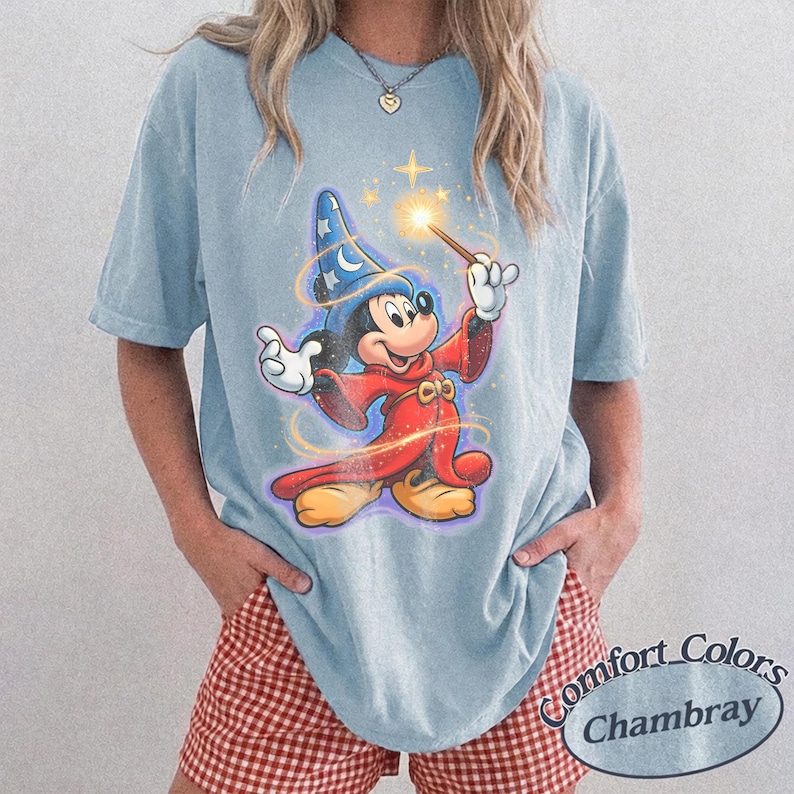 Pu&ograve; includere: T-shirt azzurra con una grafica di Topolino come mago, che tiene in mano una bacchetta con la punta luminosa. Il design include stelle e un cappello da mago blu. La maglietta ha le parole "Comfort Colors Chambray" stampate in basso a destra.