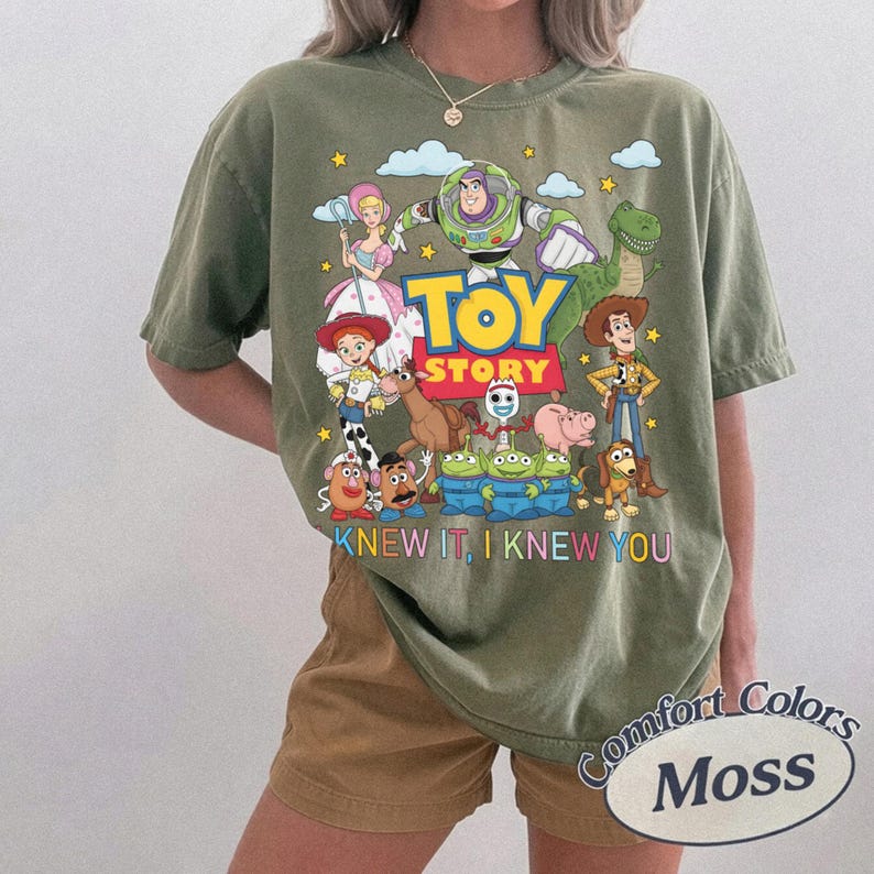 May include: An olive green short-sleeve t-shirt featuring a colorful graphic print of Toy Story characters including Buzz Lightyear, Woody, and Jessie. The text reads TOY STORY and KNEW IT, I KNEW YOU. The shirt has a crew neckline and a relaxed fit.