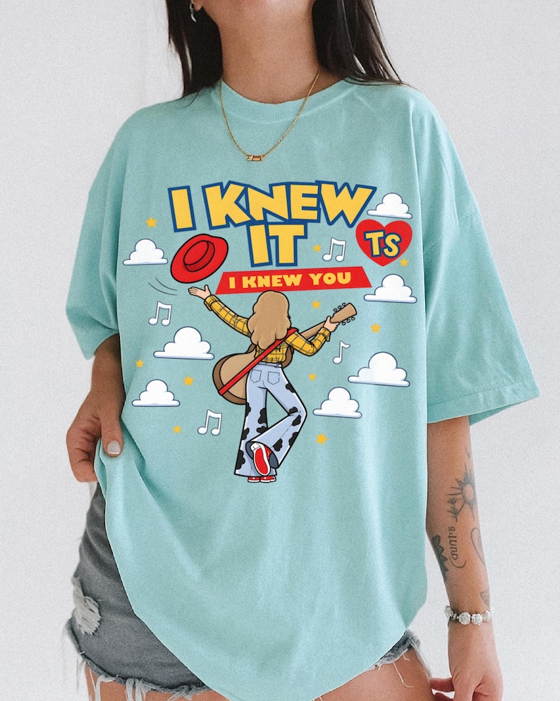 May include: A light blue t-shirt featuring a graphic of a person with long hair playing an acoustic guitar. The text reads I KNEW IT and I KNEW YOU. The design includes a red hat, a red heart with TS, white clouds, musical notes, and yellow stars.