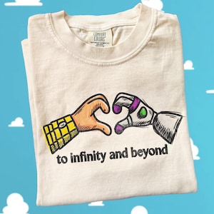 May include: A cream-colored t-shirt featuring a graphic of two hands forming a heart shape. One hand has a yellow plaid sleeve, and the other is a white and purple robotic hand. The text "to infinity and beyond" is printed in black below the hands.