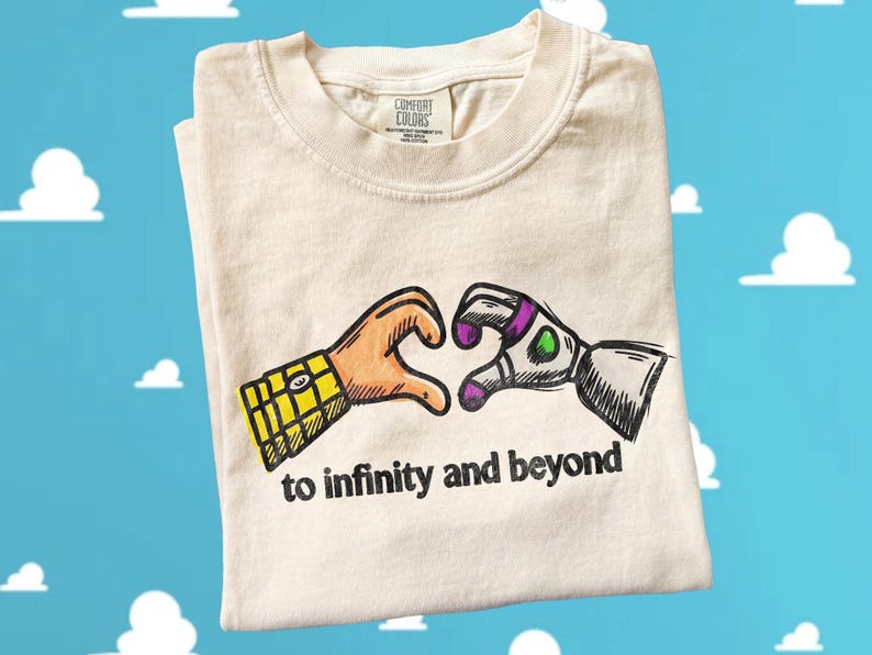 May include: A cream-colored t-shirt featuring a graphic of two hands forming a heart shape. One hand has a yellow plaid sleeve, and the other is a white and purple robotic hand. The text "to infinity and beyond" is printed in black below the hands.