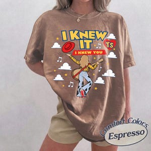 May include: A brown t-shirt featuring a graphic print of a person with long hair playing a guitar, wearing a yellow plaid shirt and cow-print trousers. The text reads I KNEW IT I KNEW YOU, accompanied by a red hat, a red heart, clouds, and musical notes.