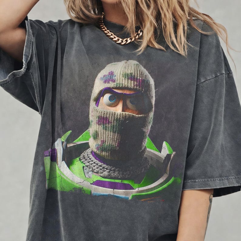 May include: A charcoal gray short-sleeve t-shirt featuring a graphic print of a character wearing a camouflage knit balaclava and a thick silver chain. The character has large blue eyes and is wearing a green and purple space-themed suit.