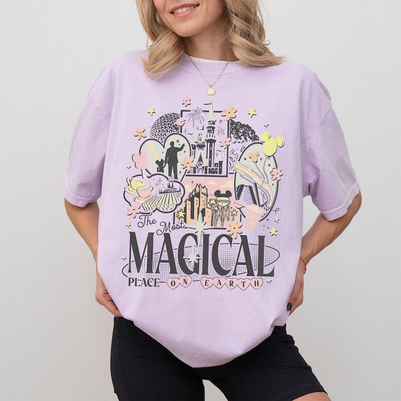 May include: A lilac t-shirt with a graphic that reads "The Most Magical Place on Earth." The design includes a silhouette, a castle, and other theme park images. The t-shirt has a loose fit.