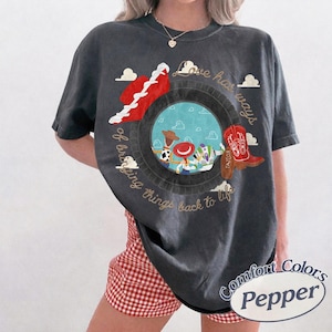 May include: A charcoal gray short-sleeve t-shirt featuring a circular graphic with a blue sky and cloud background. Inside the circle are three cartoon characters. The text reads Love has ways of bringing things back to life in gold script.