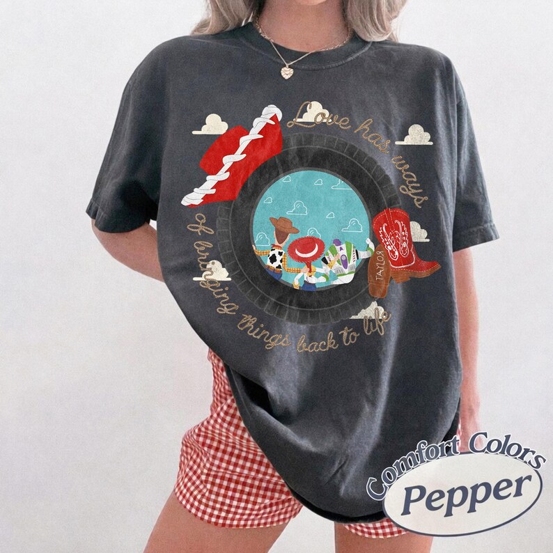 May include: A charcoal gray short-sleeve t-shirt featuring a circular graphic with a blue sky and cloud background. Inside the circle are three cartoon characters. The text reads Love has ways of bringing things back to life in gold script.