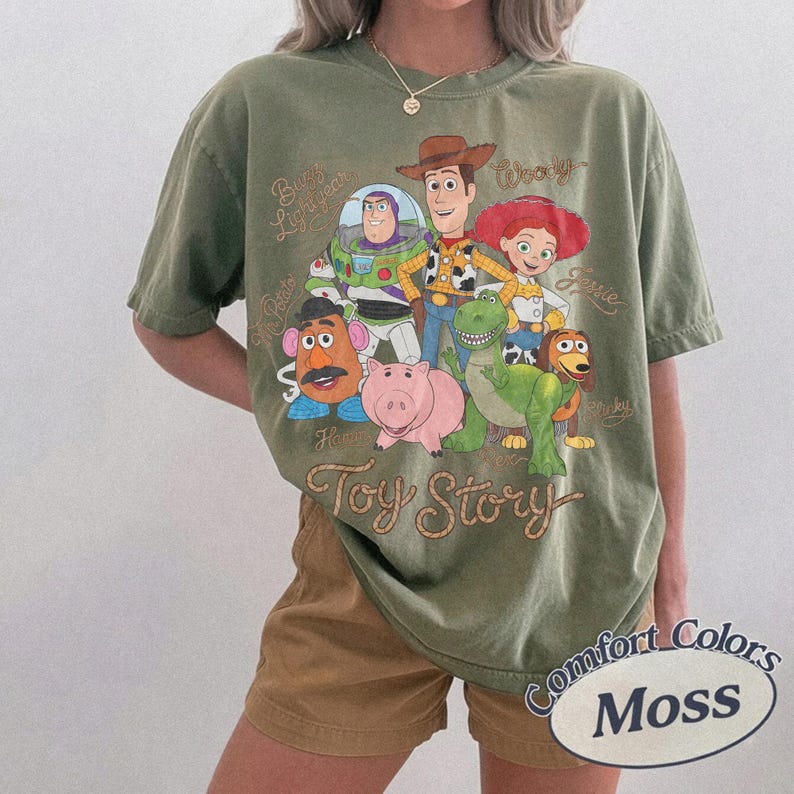 May include: An olive green short-sleeve t-shirt featuring a graphic print of Toy Story characters including Woody, Buzz Lightyear, Jessie, Mr. Potato Head, Hamm, Rex, and Slinky Dog. The text Toy Story is printed in a cursive font below the characters.