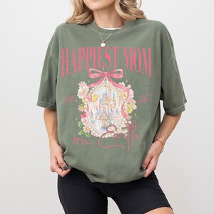 May include: Olive green t-shirt with the text "Happiest Mom on Earth" in pink. The shirt features a graphic of a castle inside a pink frame with floral accents. The shirt is a relaxed fit.
