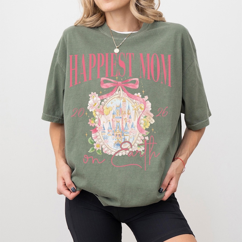 May include: Olive green t-shirt with the text "Happiest Mom on Earth" in pink. The shirt features a graphic of a castle inside a pink frame with floral accents. The shirt is a relaxed fit.