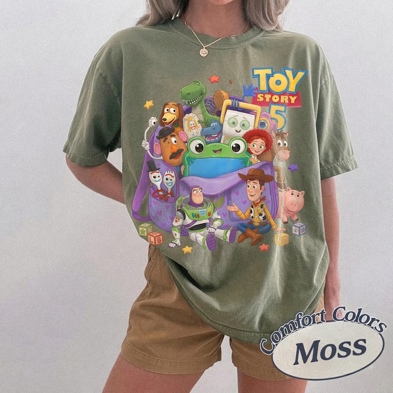 May include: An olive green short-sleeve t-shirt featuring a colorful graphic print of various Toy Story characters, including Woody, Buzz Lightyear, and a large green frog, with the text Toy Story 5 printed in blue and yellow at the top right.
