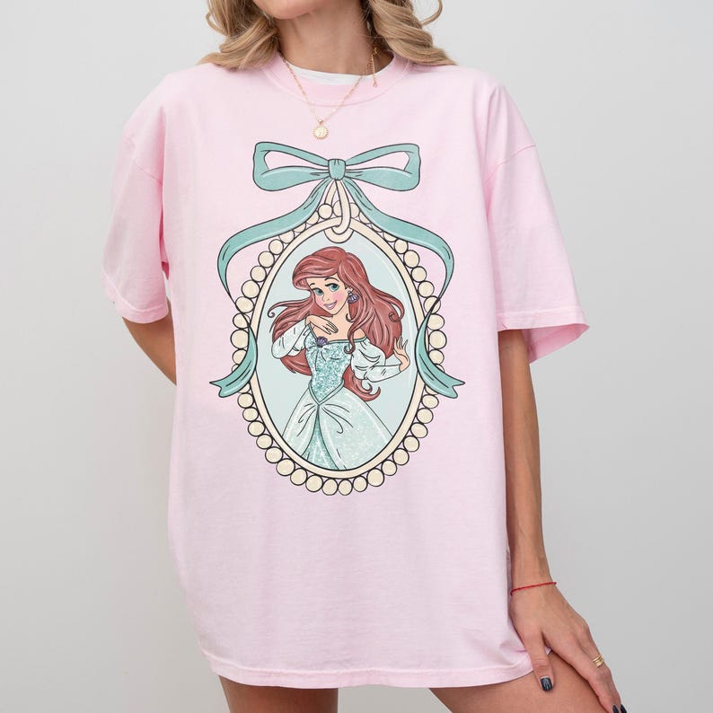 May include: A light pink t-shirt featuring a framed illustration of a cartoon character with red hair and a blue dress. The frame is surrounded by a pearl-like border and a light blue ribbon. The shirt is a casual style.