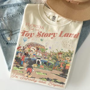 Peut inclure: T-shirt cr&egrave;me avec un graphisme de Toy Story Land en Floride. Le t-shirt pr&eacute;sente Mickey Mouse, Woody, Buzz Lightyear et d'autres personnages. Le texte comprend "Florida Est. 2018" et "To Infinity and Beyond."