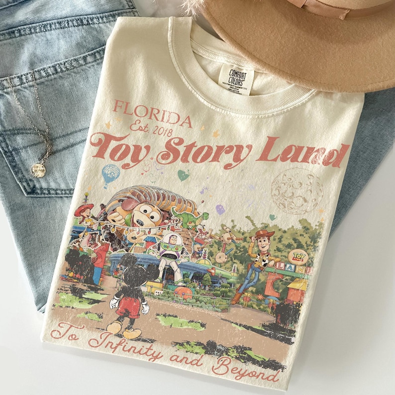Peut inclure: T-shirt cr&egrave;me avec un graphisme de Toy Story Land en Floride. Le t-shirt pr&eacute;sente Mickey Mouse, Woody, Buzz Lightyear et d'autres personnages. Le texte comprend "Florida Est. 2018" et "To Infinity and Beyond."