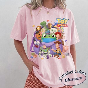 May include: A light pink t-shirt featuring a graphic print of various Toy Story characters including Woody, Buzz Lightyear, and a green frog emerging from a purple backpack. The text Toy Story 5 is printed in blue and yellow above the characters.