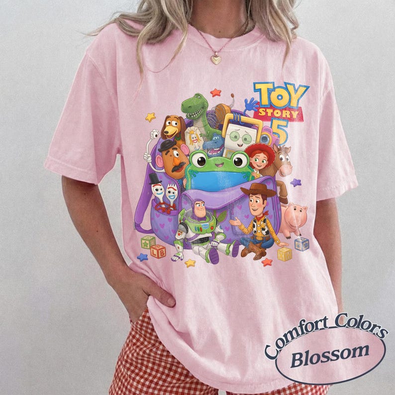 May include: A light pink t-shirt featuring a graphic print of various Toy Story characters including Woody, Buzz Lightyear, and a green frog emerging from a purple backpack. The text Toy Story 5 is printed in blue and yellow above the characters.