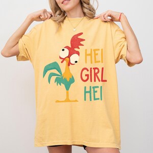 Peut inclure: T-shirt jaune moutarde avec un dessin de coq de dessin anim&eacute; et le texte "HEI GIRL HEI" en rouge, jaune et vert. Le coq a des d&eacute;tails rouges, blancs, jaunes et bleus. Le t-shirt est ample.