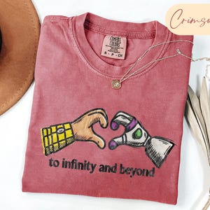 May include: A crimson short-sleeved t-shirt featuring a graphic of two hands forming a heart shape, one with a yellow plaid pattern and the other with a white, green, and purple robotic design. The text "to infinity and beyond" is printed below.