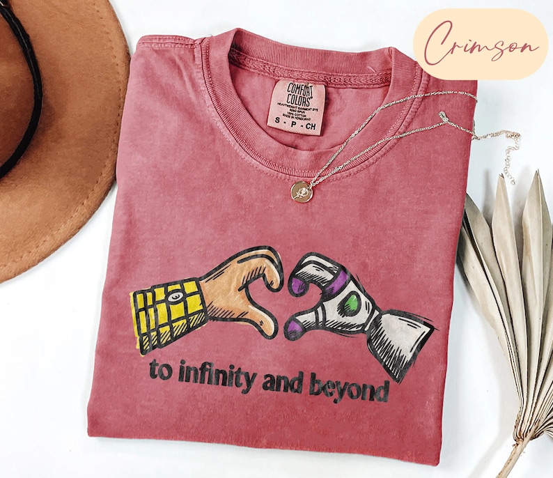 May include: A crimson short-sleeved t-shirt featuring a graphic of two hands forming a heart shape, one with a yellow plaid pattern and the other with a white, green, and purple robotic design. The text "to infinity and beyond" is printed below.