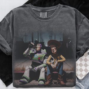 May include: Dark grey long-sleeve shirt with a graphic of Buzz Lightyear and Woody from Toy Story. Buzz is drinking from a bottle, and Woody is sitting beside him. The image has a distressed, vintage appearance.