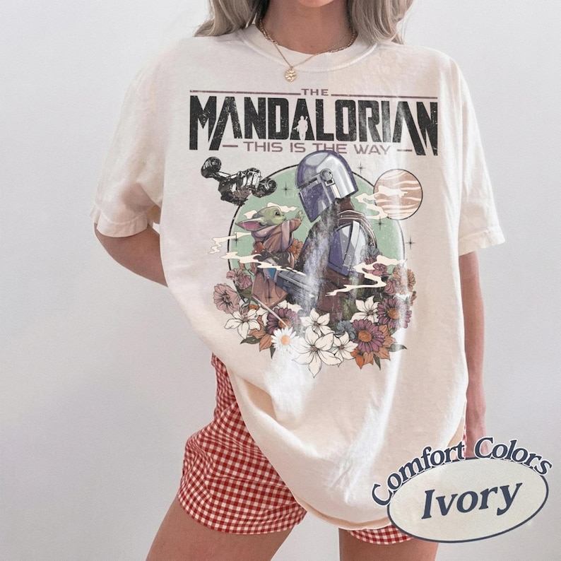 Może przedstawiać: Kremowa koszulka z grafiką The Mandalorian i Grogu otoczonych kwiatami. Tekst "THE MANDALORIAN - THIS IS THE WAY" jest wydrukowany nad obrazem. Koszulka jest marki Comfort Colors.