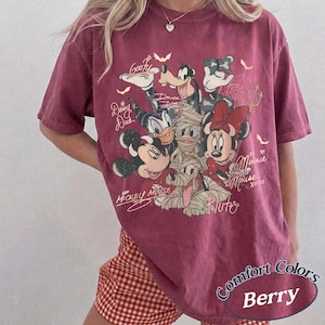 May include: Berry-coloured t-shirt with a graphic of Disney characters, including Mickey Mouse, Minnie Mouse, and Goofy. The shirt has a vintage look with the words "Mickey Mouse" and "Minnie Mouse" in script. The shirt is labelled "Comfort Colors Berry".