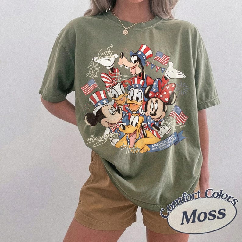 Puede incluir: Camiseta verde oliva con un dise&ntilde;o de personajes de Disney, incluyendo Mickey y Minnie, con atuendos patri&oacute;ticos. El dise&ntilde;o incluye banderas americanas y el texto "America 250 USA Anniversary 1776-2026". La camiseta est&aacute; etiquetada como "Comfort Colors Moss".