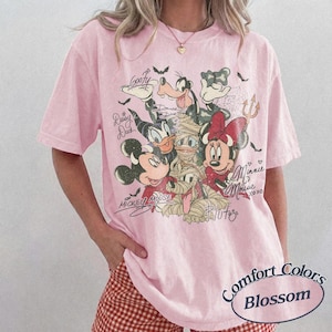 May include: Pale pink Comfort Colors Blossom t-shirt with a Halloween-themed Disney character graphic. Mickey, Minnie, Donald, Goofy, and Pluto are depicted in costume, with their signatures. The tee has short sleeves and a relaxed fit.