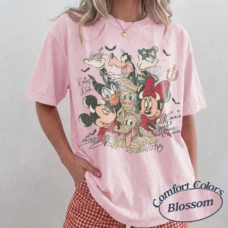 May include: Pale pink Comfort Colors Blossom t-shirt with a Halloween-themed Disney character graphic. Mickey, Minnie, Donald, Goofy, and Pluto are depicted in costume, with their signatures. The tee has short sleeves and a relaxed fit.