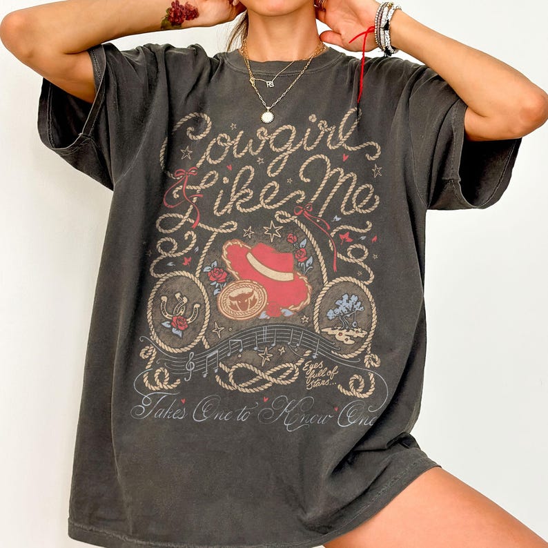Puede incluir: Una camiseta de manga corta gris carb&oacute;n con un estampado gr&aacute;fico que incluye el texto Cowgirl Like Me, Eyes full of Stars y Takes One to Know One. El dise&ntilde;o presenta un sombrero de vaquero rojo, una herradura, una cuerda y notas.