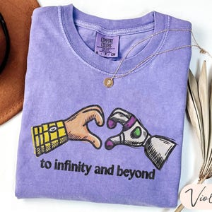 May include: A violet short-sleeved t-shirt featuring a graphic of two hands forming a heart shape, one resembling a cowboy sleeve and the other a space ranger glove. Below the graphic, the text reads to infinity and beyond in black letters.