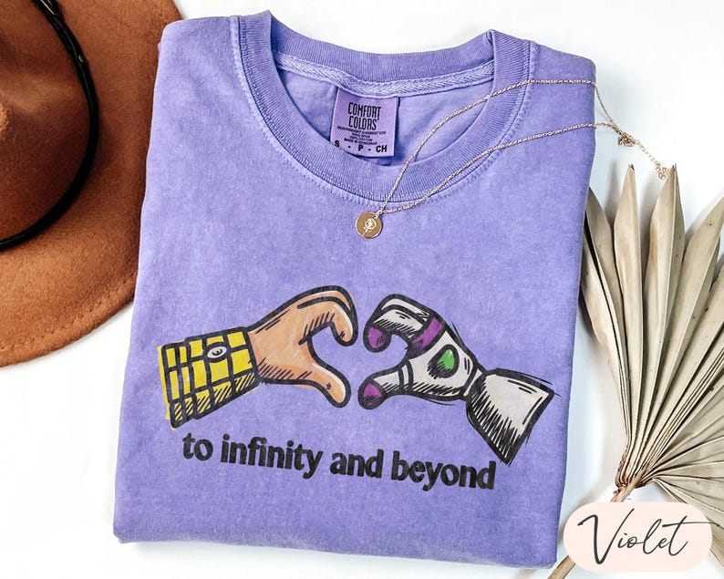 May include: A violet short-sleeved t-shirt featuring a graphic of two hands forming a heart shape, one resembling a cowboy sleeve and the other a space ranger glove. Below the graphic, the text reads to infinity and beyond in black letters.