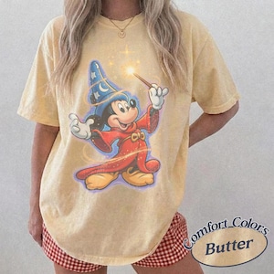 Pu&ograve; includere: T-shirt color burro con una grafica di Topolino come mago, che tiene in mano una bacchetta con una stella scintillante. Il design include un cappello da mago blu e una veste rossa. La maglietta ha le parole "Comfort Colors Butter" in basso a destra.