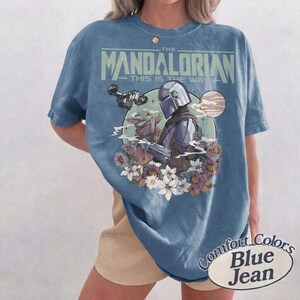 Może przedstawiać: Niebieska koszulka z grafiką The Mandalorian i Grogu otoczonych kwiatami. Tekst "THE MANDALORIAN" i "THIS IS THE WAY" jest wydrukowany nad obrazem. Koszulka jest oznaczona jako "Comfort Colors Blue Jean".