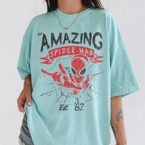 Pode incluir: Camiseta azul clara com um gr&aacute;fico desgastado do Homem-Aranha. O design inclui o texto "The Amazing Spider-Man" e "Est. '62" em vermelho e preto. A camiseta tem mangas curtas.