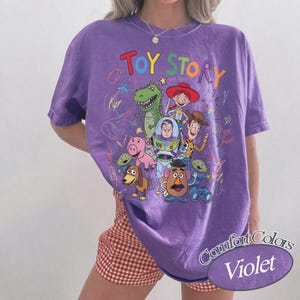 May include: A violet Comfort Colors t-shirt featuring a colorful graphic of Toy Story characters. The design includes the words "Toy Story" in a playful font, along with images of Woody, Buzz Lightyear, and other characters.