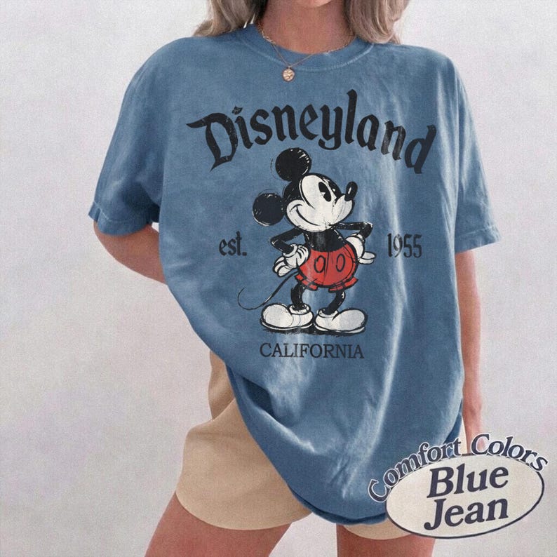 Może przedstawiać: Niebieska koszulka z grafiką Myszki Miki i napisem "Disneyland" nad postacią. Koszulka zawiera r&oacute;wnież tekst "est. 1955 CALIFORNIA" i "Comfort Colors Blue Jean".