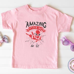 Pode incluir: Uma camiseta rosa claro com um gr&aacute;fico desgastado do Homem-Aranha. O design inclui as palavras "The Amazing Spider-Man" e "Est. '62" em preto e vermelho. A camisa tem gola redonda e mangas curtas.