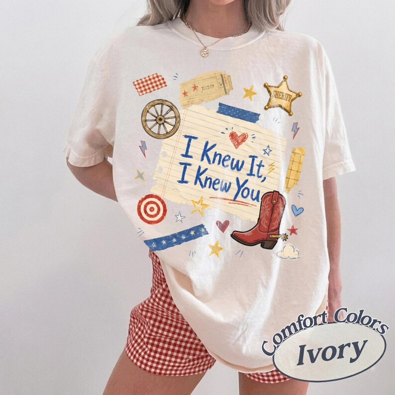 May include: An ivory t-shirt featuring a graphic print of a lined notepad with the text I Knew It, I Knew You, surrounded by illustrations of a red cowboy boot, a sheriff star, a wagon wheel, a target, and various small stars and hearts.