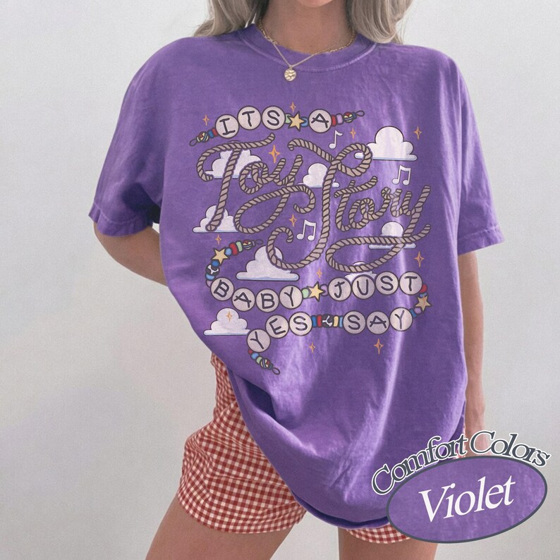May include: A violet short-sleeved t-shirt featuring a graphic print with the text "ITS A Toy Story BABY JUST YES SAY". The design includes rope-style lettering, colorful beads, small stars, musical notes, and white clouds on the front.